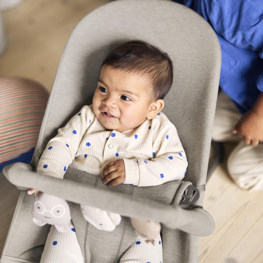 A baby in a polka dot outfit sits in a baby bouncer, while a young child in a blue shirt stands nearby, both smiling.