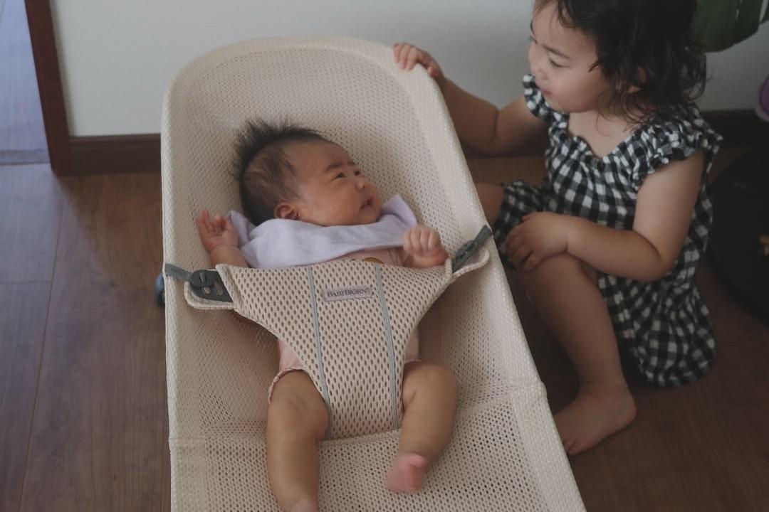 A young child in a checkered dress sits on the floor, looking fondly at a baby lying in a beige baby bouncer.