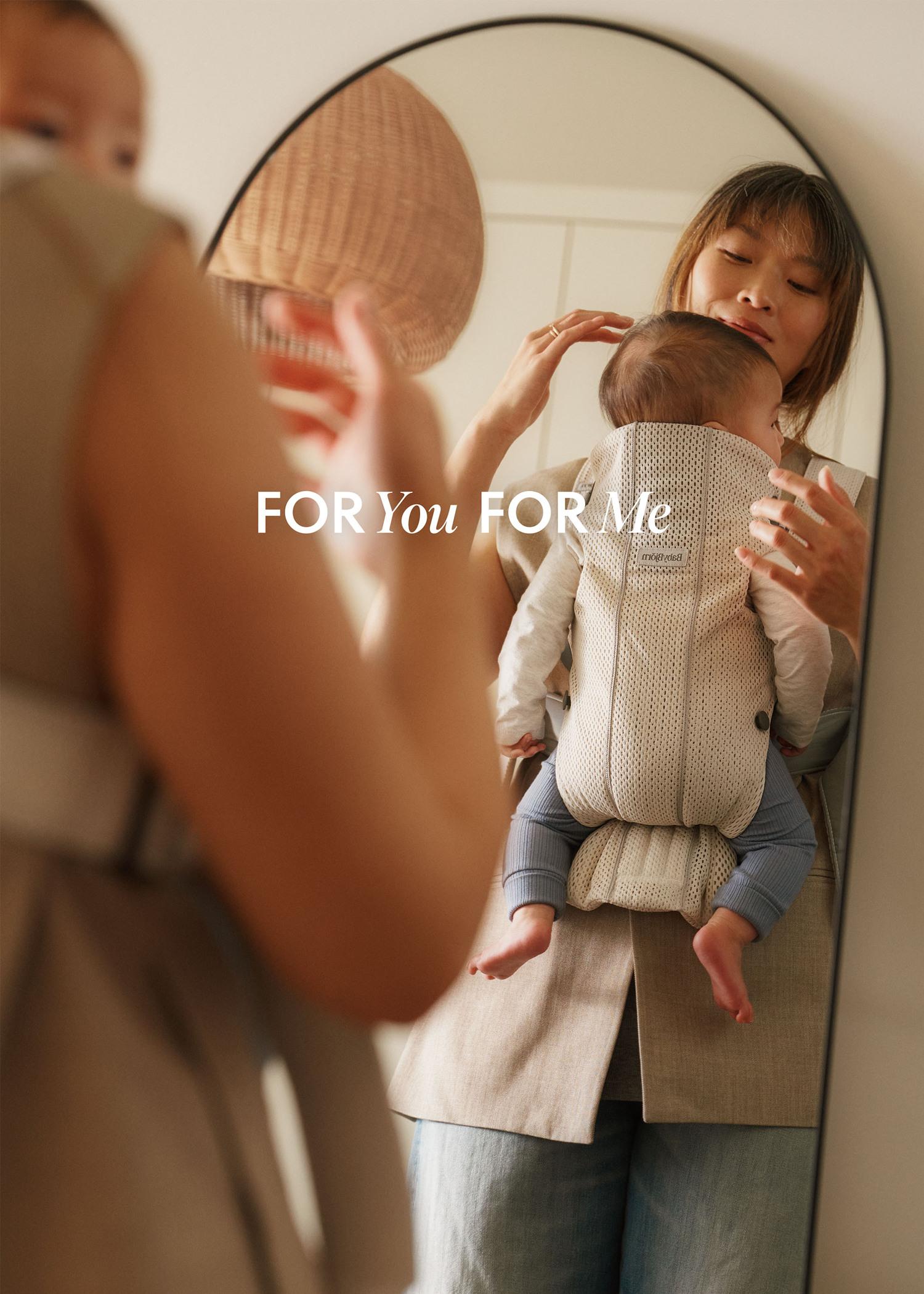 Woman in a beige outfit holding a baby in a carrier, looking in a mirror. The reflection shows her adjusting the carrier.