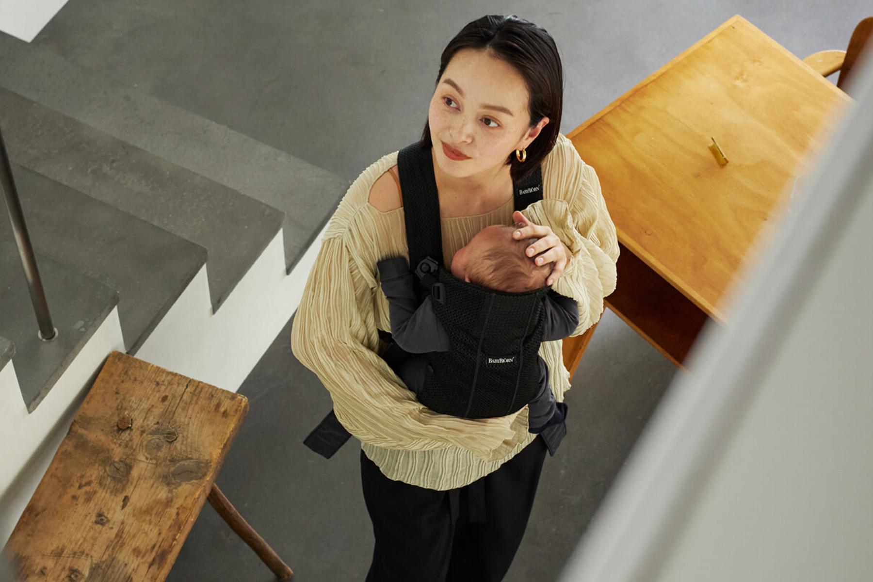 Woman carrying a baby in a black baby carrier, standing near a wooden bench and table on a gray floor.