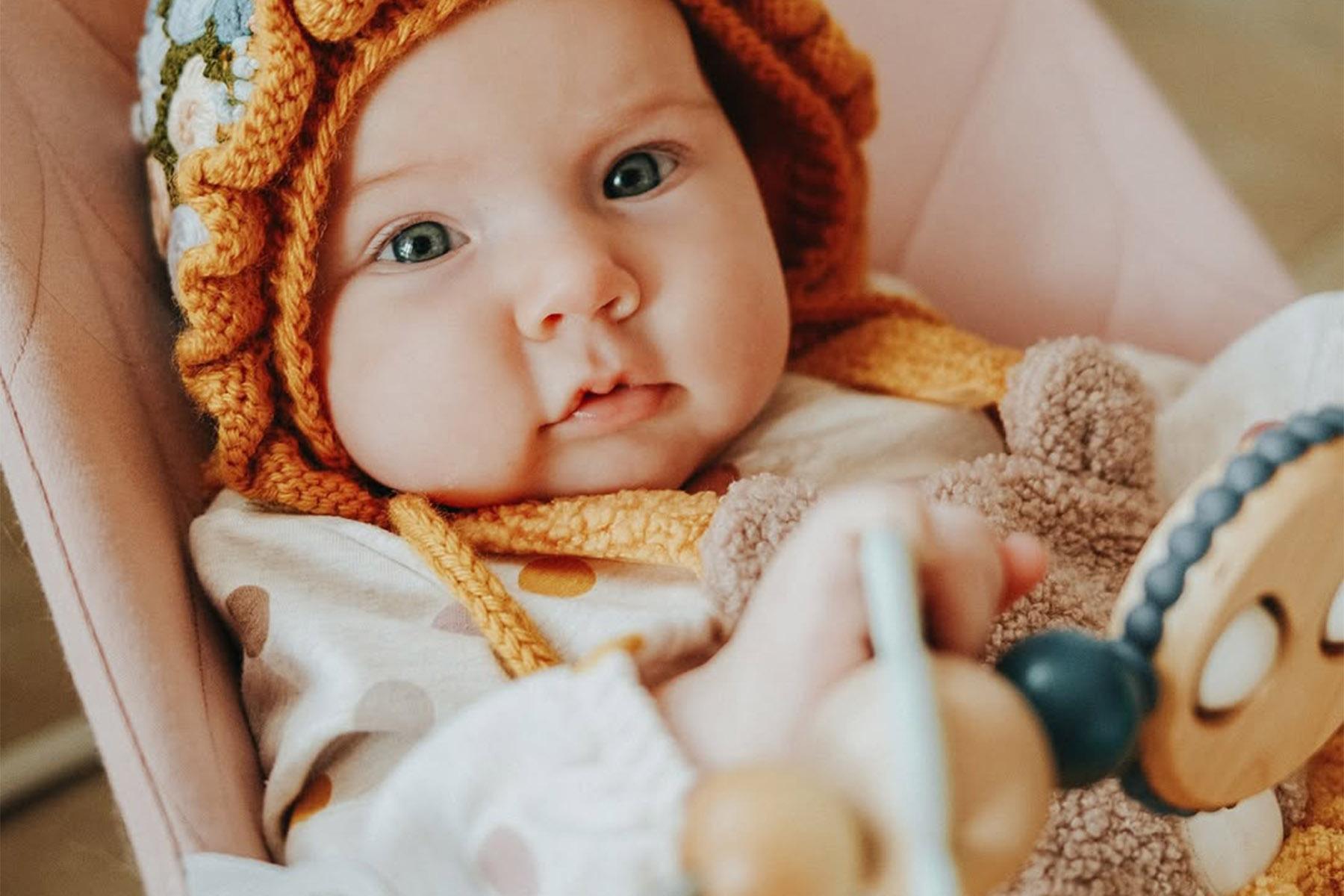 A baby wearing a knitted hat with flowers holds a wooden toy. The baby is dressed in a polka dot outfit, sitting in a pink chair.