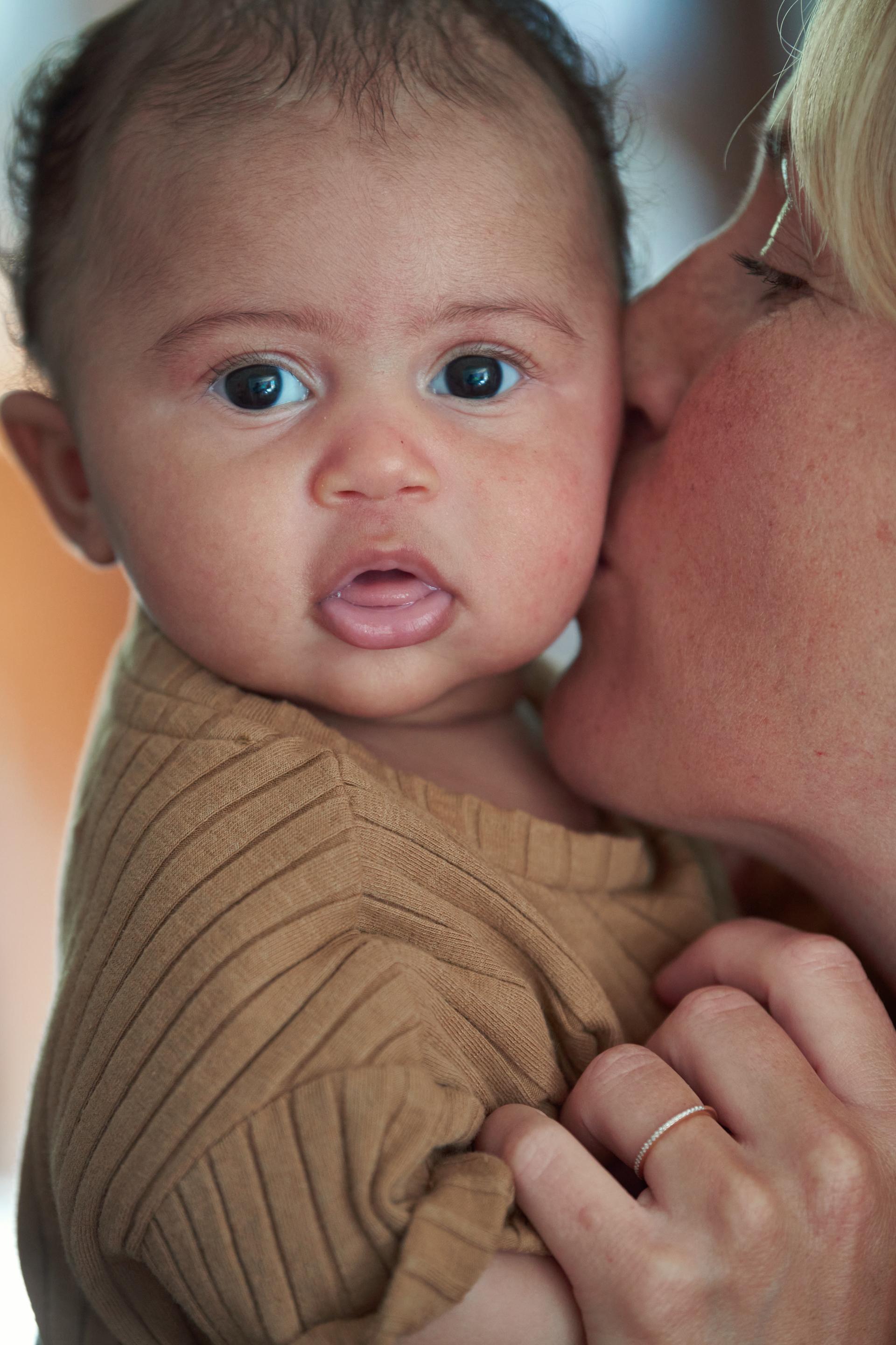 A person lovingly kisses a baby on the cheek, who is looking at the camera with wide eyes. The baby is wearing a brown outfit.