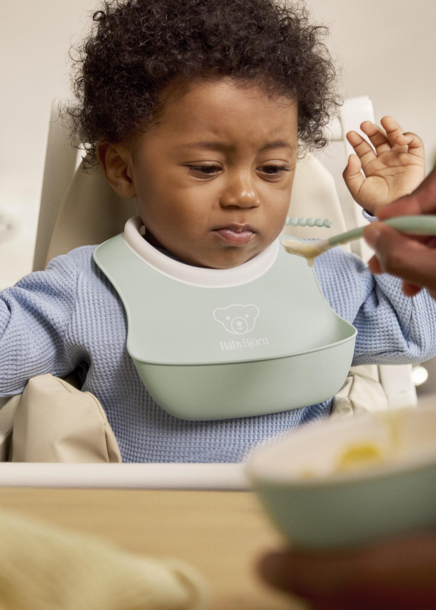 A baby in a high chair wearing a bib looks hesitant as an adult offers a spoonful of food.