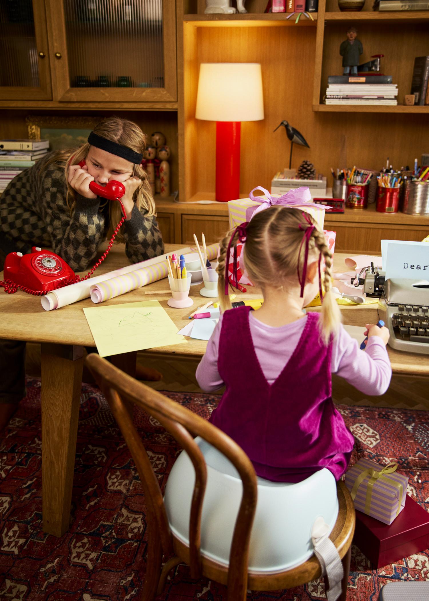 Two children in a cozy room; one talks on a red phone, the other types on a typewriter. The table is cluttered with papers and art supplies.