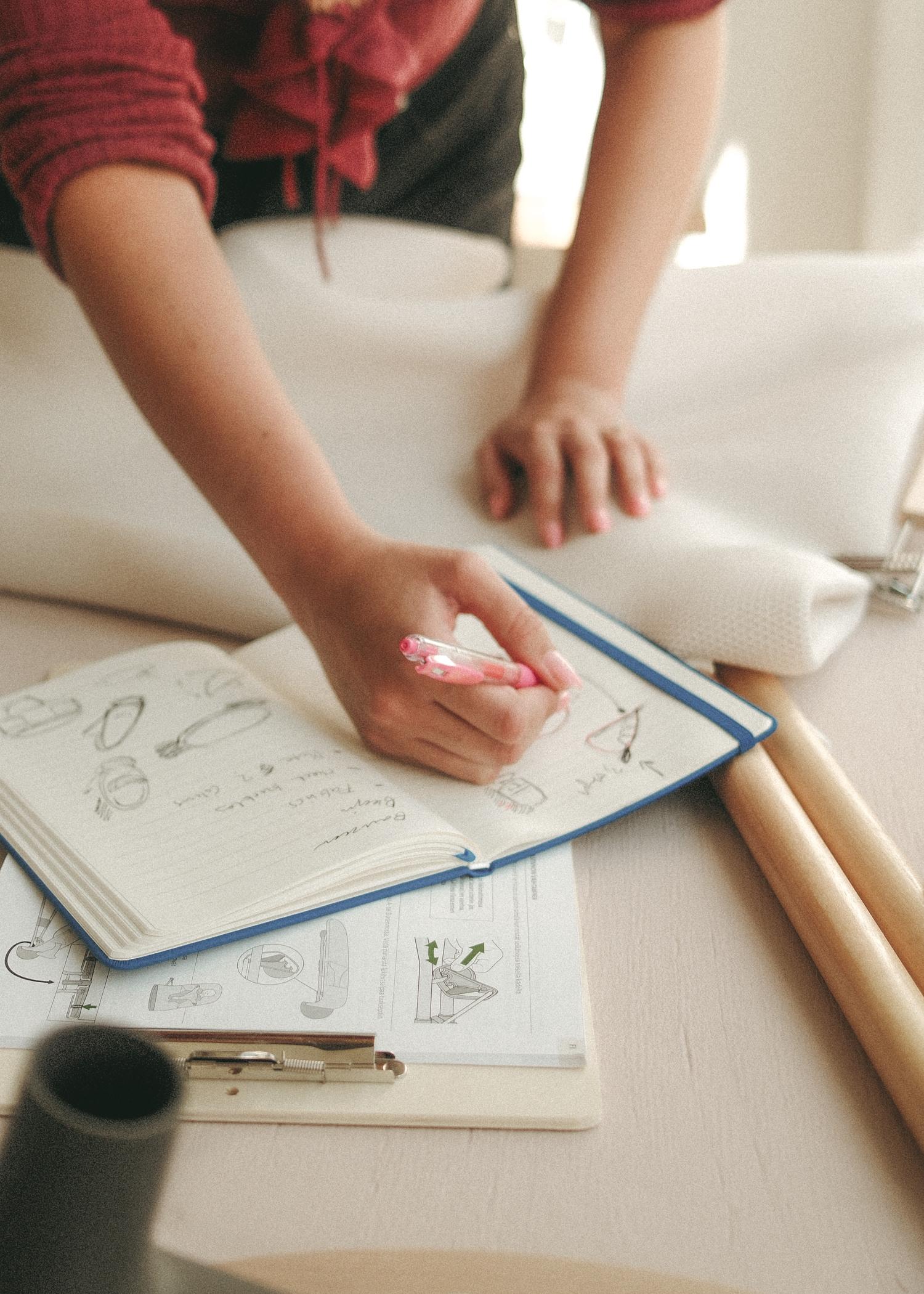 Personne dessinant dans un carnet de croquis avec des croquis et notes, à côté de divers outils créatifs sur une table claire.