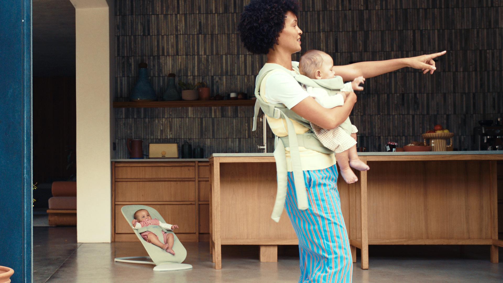 A person wearing a baby carrier points forward, while another baby sits in a bouncer nearby in a modern kitchen setting.